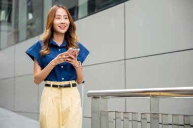 Şehir sokağında akıllı telefondan mesajlaşırken gülümseyen mavi tişörtlü genç bir kadın. İnternette arkadaşlarıyla sohbet etmek için cep telefonu uygulaması kullanan meşgul bir kız. Dışarıda güneşli bir gün.