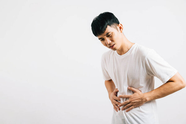 Portrait of an Asian young man experiencing stomachache and abdominal pain, suffering from digestive issues. Studio shot isolated on white background, illustrating health and medical concerns.