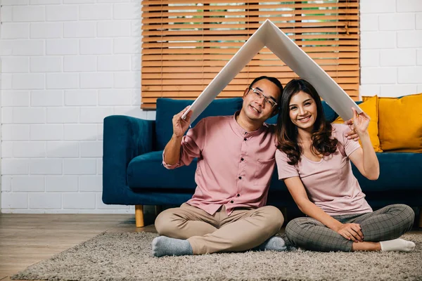 In their new home a smiling Asian couple builds a cardboard house roof signifying happiness and security. Full-length portrait against a white studio wall celebrating achievement and family bonding.
