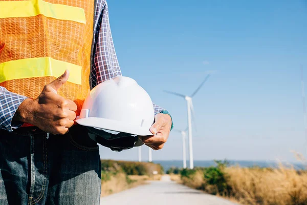 Man engineer in uniform holds helmet at wind turbine site. Signifies renewable success innovation fighting global warming. Industry leadership safety commitment illustrated.