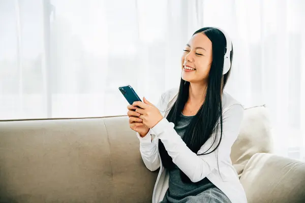 Woman with smartphone and headphones relaxes on sofa listens to music. Engaged in leisure enjoyment and technology at home. Entertainment concept.