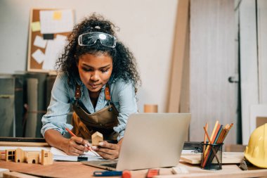 Marangoz Amerikalı siyah kadın kıvırcık saç eskizi yapıyor ve masa başında not alıyor. Ahşap marangozluk atölyesinde dizüstü bilgisayarla, genç bayan öğrenme projesi İnternet 'te marangozhanede.