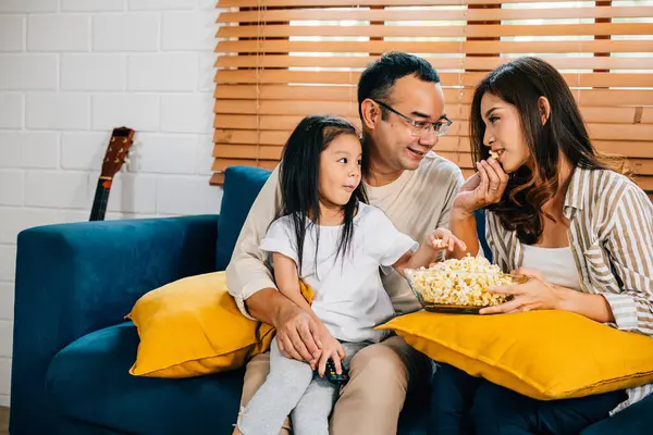 In their cozy house the family gathers on the sofa watching a video with popcorn. The father mother son daughter and schoolgirl share laughter smiles and joy epitomizing togetherness.