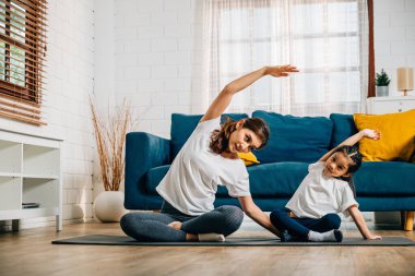 Rahat bir evde, genç bir anne ve tatlı kızı, aile yogasına hazırlanıyor. Gerinme ve denge üzerine yoğunlaşıyorlar. Gülümsemeleri, uyumlu ve neşeli anlarını yansıtıyor..