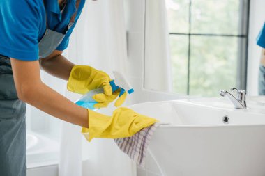 Ev işlerine odaklanmış üniformalı bir kadın banyo lavabosu ve musluğu temizliyor. Ev işlerine olan bağlılığı, evdeki saf hijyen ve parlak eşyalar üzerinde duruyor. Sprey temizleyici