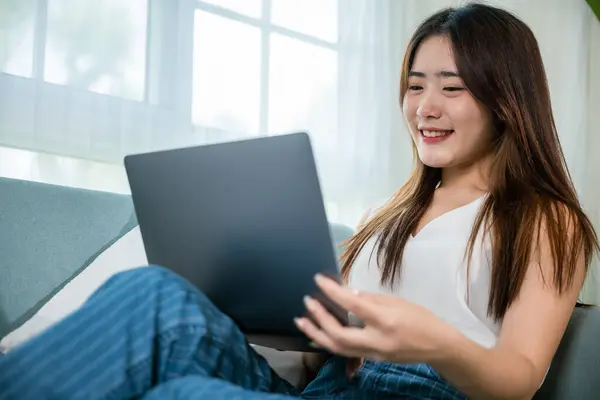 Happy woman typing email on notebook computer, Asian young female smiling sitting relaxing on sofa using laptop in living room at home, freelance browsing through the internet during free time