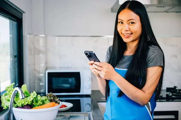 A cheerful young woman in the kitchen smiles while using her cellphone leaning on the table. Typing messages she multitasks cooking dinner in a modern fun atmosphere at home. mobile phone