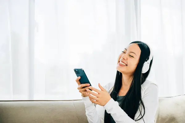 Woman relaxes on sofa listens to music on smartphone wears headphones. Engaged in leisure enjoyment and technology at home. Entertainment and relaxation concept.