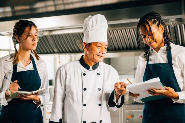 Asyalı kıdemli şef, bir restoran mutfak atölyesinde çeşitli öğrencilere yemek pişirme yöntemleri öğretiyor. Takım çalışmasına odaklan ve not al. Profesyonel eğitim. Gıda Eğitimi