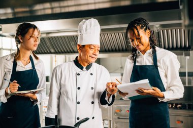 Profesyonel Asyalı kıdemli şef çeşitli öğrencilere bir restoran mutfağı atölyesinde rehberlik ediyor. Takım çalışmasına odaklan ve not al. Eğitim ortamı. Gıda Eğitimi