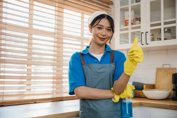 Housework-ready woman in uniform with apron and rubber glove holds spray bottle for home cleaning. Emphasizing hygiene and cleanliness. Clean disinfect home care. maid with liquid.