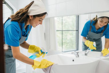 Üniformalı bir kadın banyo lavabosunu ve musluğu spreyle temizler. Ev işlerine olan bağlılığı saf hijyen ve parlak eşyalar sağlıyor. Sprey temizleyici