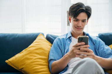 Evdeki bir kanepede gülümseyen genç bir adam telefonda konuşuyor iletişim ve teknolojiyi kucaklıyor. Onun mutluluğu başarılı yaşam ve rahatlamayı yansıtır. sosyal medyada kaydediliyor.