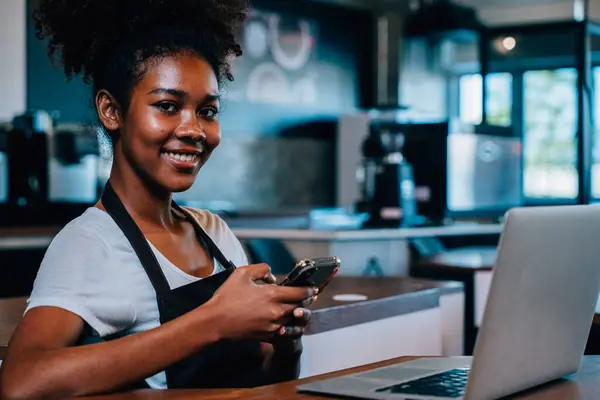 Close up portrait of concentrated black woman coffee shop owner in apron using smartphone in her cafe. Multi tasking barista manages orders communicates runs her own thriving business.