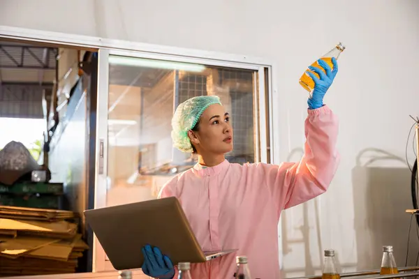 In a beverage factory a woman in a uniform checks bottles on a conveyor belt ensuring liquid quality using a laptop for meticulous quality control.