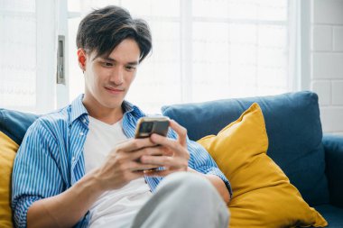 Kanepede rahat bir şekilde otururken gülümseyen ve telefonla konuşan genç bir adam. Başarılı bir iletişim ve teknolojiye sahip. Mutluluk ve rahatlama yayıyor. sosyal medyada kaydediliyor.