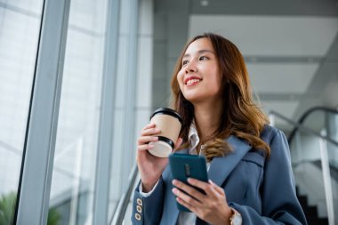 Elinde kahve fincanı ve akıllı telefonu olan mavi gömlekli genç Asyalı iş kadını. Gülümsüyor, pencereden dışarı bakıyor ve ofisteki asansörün arka planı bulanık..