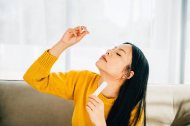 COVID-19 'un kendi kendini test ettiğini vurgulayan Asyalı kadın, evinde hızlı bir antijen test kiti kullanıyor ve burnuna çubuk sokuyor. Virüs önleme ve pandemik koruma tedbirleri.