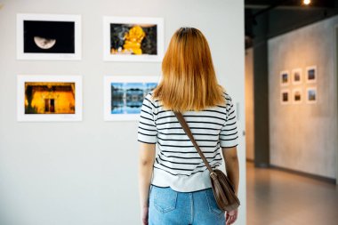 Asyalı bir kadın renkli çerçeveli resimlerin önünde dikiliyor beyaz duvarda resimler, genç bir kadın fotoğraf çerçevesinde sergi galerisine yaslanıyor,