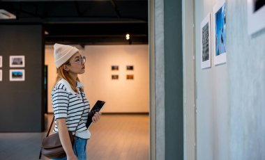 Asyalı bir kadın sanat galerisinde tablet tutuyor. Çerçeveli resimlerin önünde beyaz duvarda, fotoğraf çerçevesinde genç bir kadın sergi sanat galerisinde dijital kitap tutuyor.
