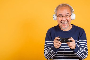 Yüzünde kulaklıkla gülümseyen Asyalı yaşlı adam oyun kumandası stüdyosu görüntüsü izole edilmiş sarı arka plan, yaşlı adam joystick konsoluyla video oyunu oynamaktan mutlu.