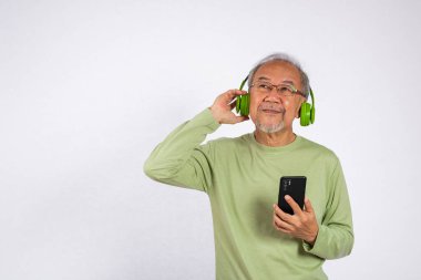 Gözlüklü ve kulaklıklı gülümseyen Asyalı yaşlı adam akıllı telefon tutuyor. Kulaklıklarda müzik dinliyor. Beyaz arka planda izole edilmiş, müziğinin tadını çıkarıyor.