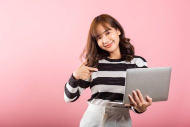 Mutlu Asyalı güzel bir kadının portresi dizüstü bilgisayarla bilgisayarı işaret ederek gülümseyen bir gülümseme, pembe arka planda izole edilmiş bir stüdyo resmi,