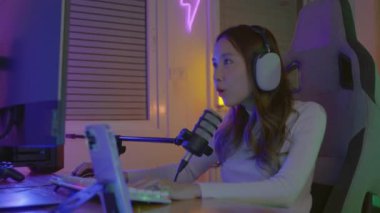 Oyun odasında mikrofonla konuşan mutlu Asyalı kadın, kulaklık takan genç oyuncu kadın bilgisayarda video oyunları oynuyor hayranlarıyla canlı yayında sohbet ediyor, oyun konsepti