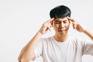 Stres ve acı dolu bir baş ağrısı yaşayan Asyalı bir adam üzgün ve yorgun bir ifadeyle başını tutar. Stüdyo çekimi beyaz arka planda izole edilmiş, onun mutsuzluğunu ve rahatsızlığını gösteriyor..