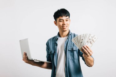 Asyalı gülümseyen genç adam dizüstü bilgisayar ve para fanatiği stüdyo görüntüsü izole beyaz arka plan, mutlu yaşam tarzı işadamı dolar banknotları içinde para tutuyor.