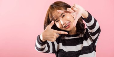 Asyalı bir fotoğrafçı, parmağıyla yüzünün önünde bir çerçeve oluştururken gülümsüyor. Stüdyo çekimi pembe, yaratıcılık ve odaklanma üzerine izole edilmiş..