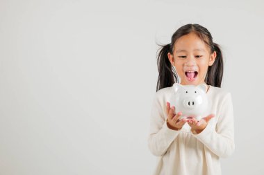 Asyalı küçük bir kız elinde domuz kumbarası, beyaz arka planda izole edilmiş stüdyoya bakıyor, mutlu bir anaokulu çocuk yaşam tarzı gülümsüyor, beyaz kumbara, Tayland kişisel para tasarrufu.