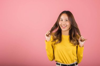 Uzun saçlı, sarı tişörtlü çekici bir Asyalı kadın gülümsüyor ve saçını arkada tutuyor. Pembe arka planda izole edilmiş, mutlu ve sevimli bir kız.