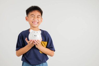 Asyalı küçük çocuk kumbarasını tutuyor ve beyaz arka planda izole edilmiş stüdyo kamerasına bakıyor, mutlu çocuk yaşam tarzı ile gülümseyen beyaz kumbara, Tayland kişisel para tasarrufu