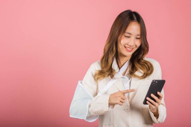 Kendine güvenen bir kadın kaza sonrası gülümseyen kırık bir kol ve tedavi için kol bandı takan ve akıllı telefon tutan mutlu Asyalı kadın kol askısı pembe arka plan, sosyal medya, kopyalama alanı izole edilmiş.