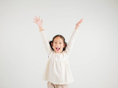 Sevinçli bir kız çocuğu başarısını kutlamak için heyecanla evet diyerek yumruklarını kaldırıyor. Beyaz arka planda çekilen mutlu bir anaokulu çocuğunun Asyalı portresi.