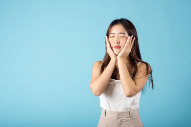 Doğal makyajlı ve sağlıklı yüzüyle gülümseyen yakın bir kadın, sağlıklı yüz derisine dokunan Asyalı genç bir kadın, mavi arka planda izole edilmiş bir çekim, cilt bakımı ve güzellik konsepti.