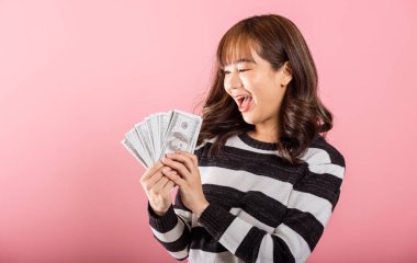 Asyalı mutlu portre gülümseyen güzel genç kadın elinde para yelpazesi banknotları pembe arka planda izole edilmiş 100 dolarlık banknotlar.