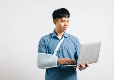 Kendine güvenen Asyalı profesyonel, kırık bir koluyla bir dizüstü bilgisayar üzerinde çalışmaya devam ediyor. Stüdyo, kazadan sonra kararlılık ve iyileşme göstergesi olarak beyaz üzerine izole edildi..