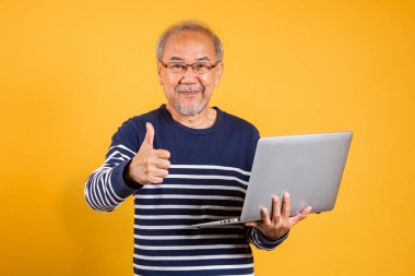 Gözlüklü Asyalı yaşlı adamın portresi dizüstü bilgisayar kullanıyor ve stüdyo fotoğrafı çekiyor izole edilmiş sarı arka plan, gülümseyen yaşlı adam gri saçlı başarı memnuniyeti kavramı.