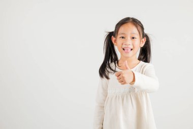 Beyaz arka planda izole edilmiş bir stüdyo portresindeki kendine güvenen Asyalı genç kız onay işareti yapıyor, anlaşma ve başarıyı simgeliyor. Anaokulu çocuk gülümsemesi pozitiflik ve memnuniyet yayar.