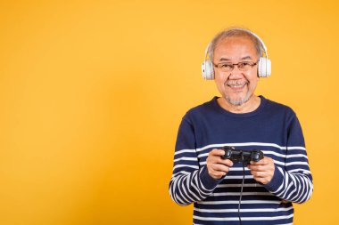 Yüzünde kulaklıkla gülümseyen Asyalı yaşlı adam oyun kumandası stüdyosu görüntüsü izole edilmiş sarı arka plan, yaşlı adam joystick konsoluyla video oyunu oynamaktan mutlu.