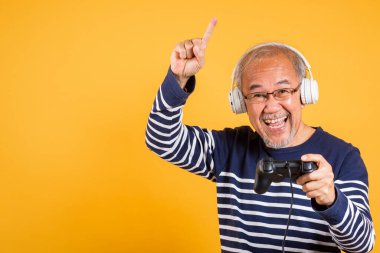 Yüzünde kulaklıkla gülümseyen Asyalı yaşlı bir adam oyun kontrol stüdyosu çekim izole sarı arka plan, son sınıf öğrencisi oyun konsolu ile bir video oyunu oynadığını hissediyor