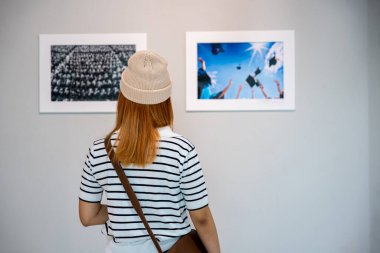 Asian young woman standing she looking art gallery in front of colorful framed paintings pictures on white wall, female watch at photo frame to leaning against at exhibit museum, Back view