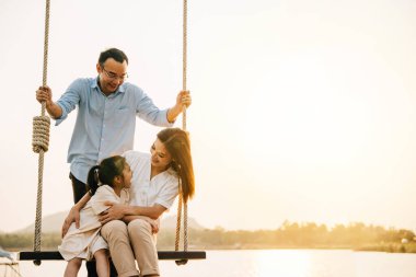 Anne ve baba ve kızı birlikte salıncakta sallanırken anne ve oğlu arka planda oturup mutlu ve neşeli bir aile aktivitesi izliyorlar.