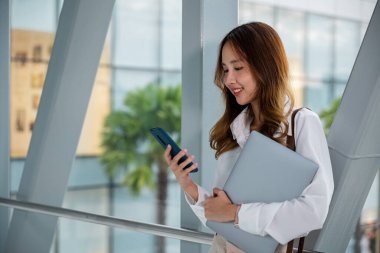 Beyaz gömlekli profesyonel bir kadın havaalanında dizüstü bilgisayar ve telefonla birden fazla iş yapıyor. Üretken bir çalışma yürütülüyor. Seyahat ederken bağlı ve üretken kalın