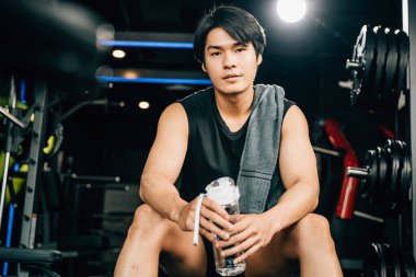 Asyalı genç adam portresi siyah spor kıyafetleri içinde dinlenmek için oturmuş spor salonunda su ya da protein tutarken ağırlık antrenmanı, sağlıklı yaşam tarzı konsepti.