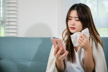 Asian young woman sick sitting down on sofa with cold blowing her nose and checking smart mobile phone to cell to doctor online, depressed sad sick female holding tissues and smartphone in living room