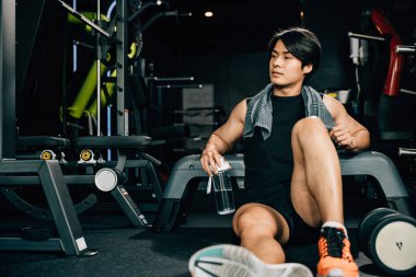 Spor kıyafetleriyle eğitilmiş Asyalı bir gencin portresi spor salonunda antrenman, ağırlık eğitimi, sağlıklı yaşam tarzı konsepti sonrasında elinde su ya da proteinle bankta dinleniyor.