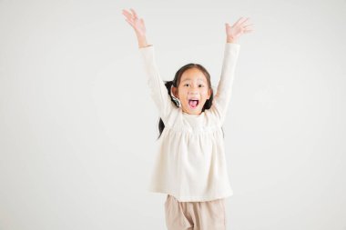 Sevinçli bir kız çocuğu başarısını kutlamak için heyecanla evet diyerek yumruklarını kaldırıyor. Beyaz arka planda çekilen mutlu bir anaokulu çocuğunun Asyalı portresi.